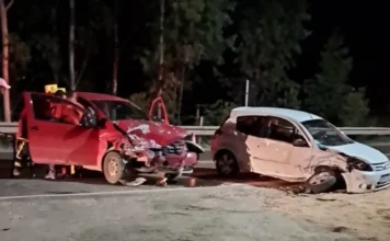 Acidente em Lauro Müller deixa seis feridos no Guatá Fotografia noturna em uma estrada de asfalto mostrando dois carros de passeio, um Volkswagen Fox vermelho e um Ford Ka branco, com danos severos na parte frontal após uma colisão. O Fox vermelho está à esquerda, com a porta do motorista aberta e socorristas dos bombeiros ao redor. O Ford Ka branco está à direita, posicionado diagonalmente na pista. O cenário é iluminado pelos faróis das viaturas e há vegetação escura ao fundo.