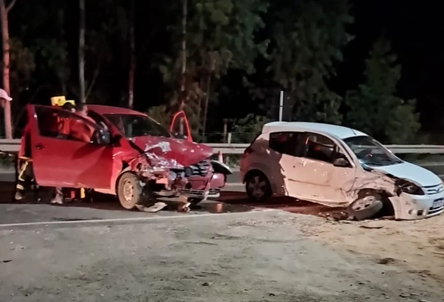 Fotografia noturna em uma estrada de asfalto mostrando dois carros de passeio, um Volkswagen Fox vermelho e um Ford Ka branco, com danos severos na parte frontal após uma colisão. O Fox vermelho está à esquerda, com a porta do motorista aberta e socorristas dos bombeiros ao redor. O Ford Ka branco está à direita, posicionado diagonalmente na pista. O cenário é iluminado pelos faróis das viaturas e há vegetação escura ao fundo.