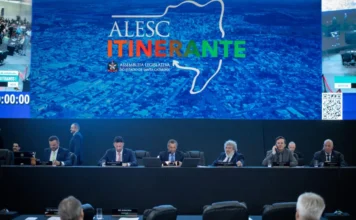 Alesc aprova 15 projetos em sessão itinerante em Curitibanos Sessão plenária da Alesc Itinerante em Curitibanos. Deputados estaduais catarinenses sentados em uma mesa de conferência principal, com um grande telão de vídeo ao fundo exibindo o mapa de Santa Catarina e o logotipo da Alesc Itinerante. A imagem mostra o auditório lotado com a plateia de costas e a mesa principal bem iluminada. Foco na Assembleia Legislativa do Estado de Santa Catarina e deputados catarinenses.
