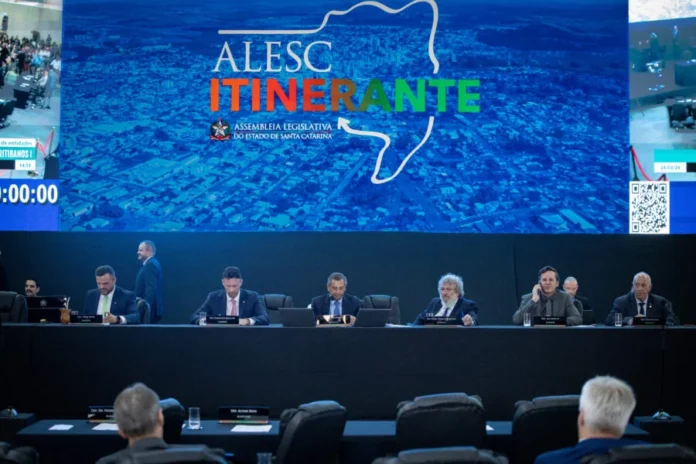 Alesc aprova 15 projetos em sessão itinerante em Curitibanos Sessão plenária da Alesc Itinerante em Curitibanos. Deputados estaduais catarinenses sentados em uma mesa de conferência principal, com um grande telão de vídeo ao fundo exibindo o mapa de Santa Catarina e o logotipo da Alesc Itinerante. A imagem mostra o auditório lotado com a plateia de costas e a mesa principal bem iluminada. Foco na Assembleia Legislativa do Estado de Santa Catarina e deputados catarinenses.