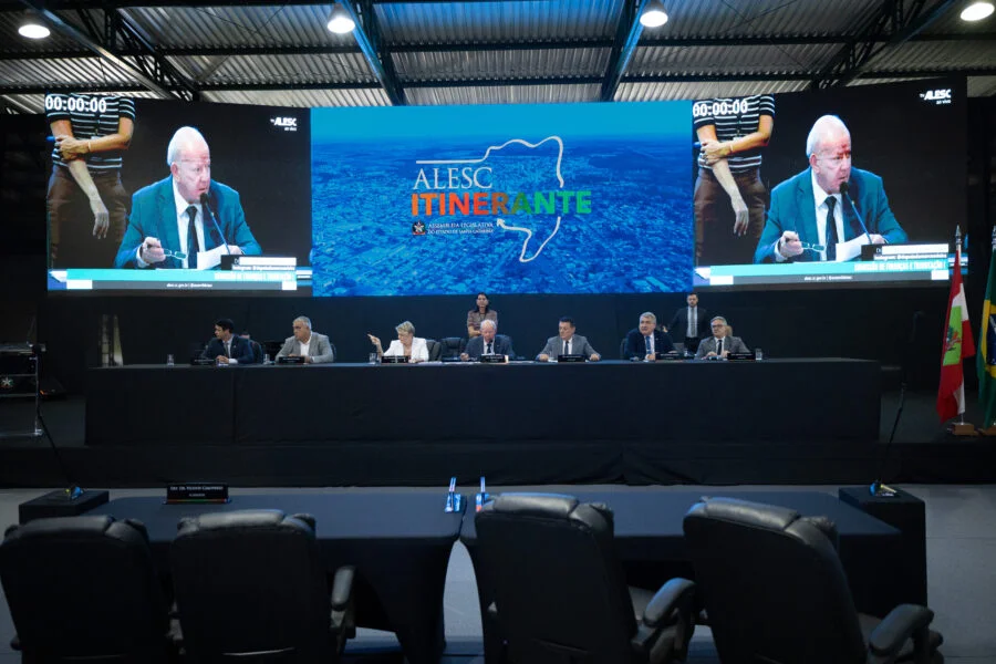 Reunião da Comissão de Finanças e Tributação da Alesc Itinerante em Curitibanos, Santa Catarina, com deputados estaduais em mesa de debate e telão de LED ao fundo exibindo a logo da Assembleia Legislativa do Estado de Santa Catarina.