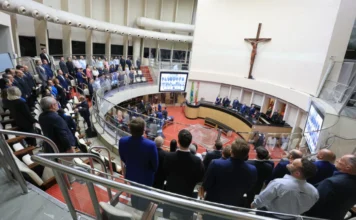 Alesc celebra 68 anos do Crea-SC em sessão especial Vista de ângulo alto do Plenário da Alesc lotado durante uma sessão especial de homenagem aos 68 anos do Crea-SC. O Coral da Alesc e os presentes estão de pé para o Hino Nacional. A cena mostra a mesa principal com autoridades, o grande crucifixo e telas de vídeo.