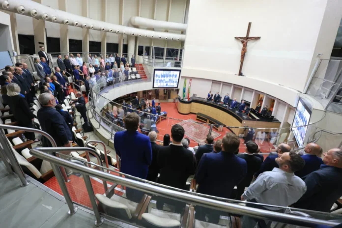 Alesc celebra 68 anos do Crea-SC em sessão especial Vista de ângulo alto do Plenário da Alesc lotado durante uma sessão especial de homenagem aos 68 anos do Crea-SC. O Coral da Alesc e os presentes estão de pé para o Hino Nacional. A cena mostra a mesa principal com autoridades, o grande crucifixo e telas de vídeo.