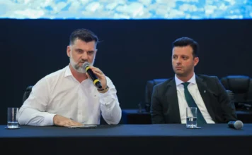 Alesc cobra cronograma para obras na Serra do Corvo Branco Prefeitos de Urubici e Grão-Pará cobram agilidade nas obras da SC-370 durante reunião da Alesc Itinerante. O prefeito Leandro de Souza Corrêa (direita), de camisa branca, fala assertivamente ao microfone sobre a pavimentação na Serra do Corvo Branco, enquanto o prefeito Hélio Alberton Júnior (esquerda), de terno e gravata, ouve atentamente. A cena ocorre em um ambiente formal com mesa coberta e painel de LED ao fundo.