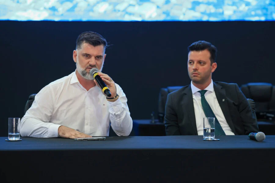 Alesc cobra cronograma para obras na Serra do Corvo Branco Prefeitos de Urubici e Grão-Pará cobram agilidade nas obras da SC-370 durante reunião da Alesc Itinerante. O prefeito Leandro de Souza Corrêa (direita), de camisa branca, fala assertivamente ao microfone sobre a pavimentação na Serra do Corvo Branco, enquanto o prefeito Hélio Alberton Júnior (esquerda), de terno e gravata, ouve atentamente. A cena ocorre em um ambiente formal com mesa coberta e painel de LED ao fundo.