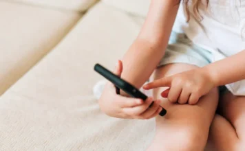 Alesc debate vício em telas e saúde mental infantil Criança pequena sentada em um sofá claro segura e toca na tela de um smartphone preto. A imagem ilustra a alta exposição de crianças a telas e dispositivos móveis.