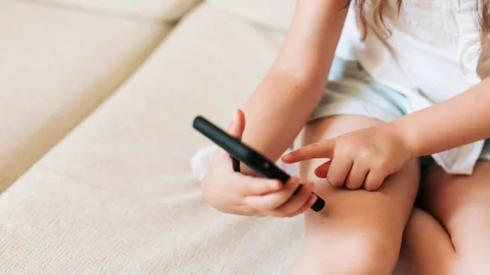 Alesc debate vício em telas e saúde mental infantil Criança pequena sentada em um sofá claro segura e toca na tela de um smartphone preto. A imagem ilustra a alta exposição de crianças a telas e dispositivos móveis.