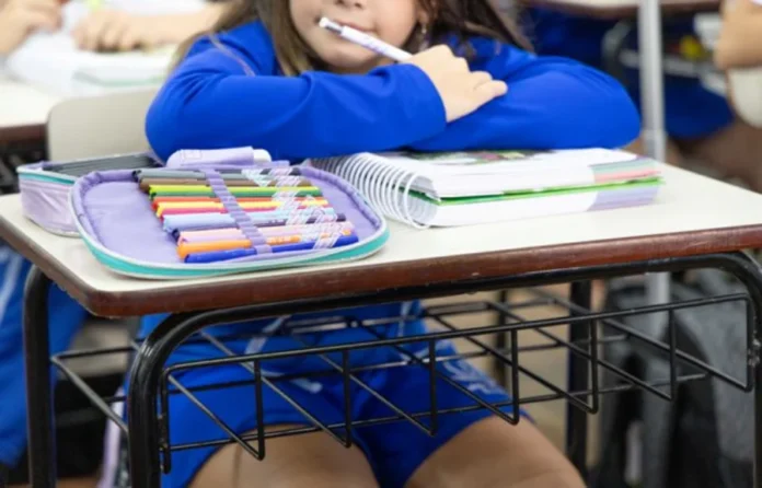 Alesc lança Prêmio Paz nas Escolas para estudantes de SC Fotografia em plano médio e foco seletivo mostrando uma estudante sentada em uma carteira escolar. A aluna veste um uniforme azul e está com os braços apoiados sobre a mesa, segurando uma caneta branca próxima ao rosto. À sua frente, destacam-se um caderno espiral branco e um estojo aberto exibindo uma fileira de canetas hidrográficas coloridas. O ambiente sugere uma sala de aula iluminada.