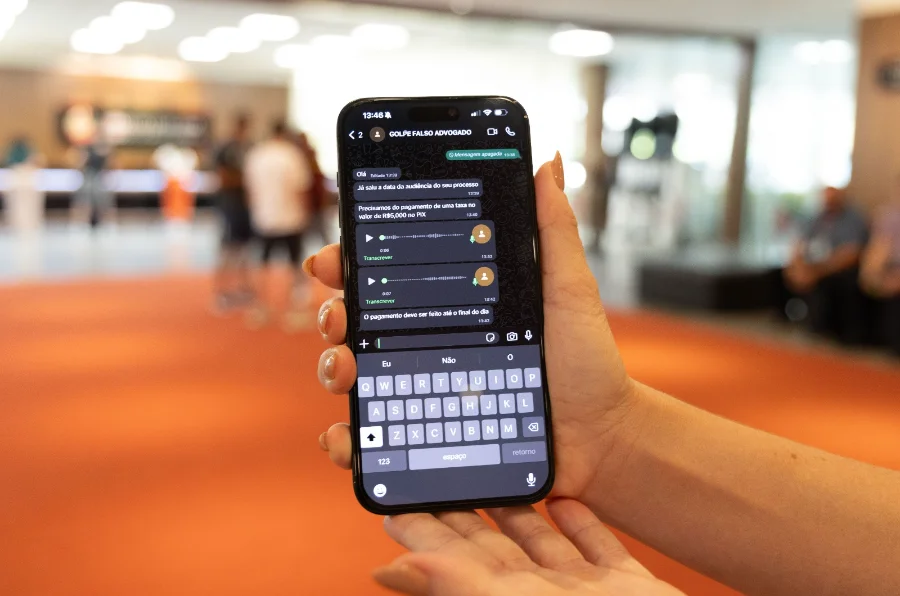 Close-up de uma mão segurando um smartphone em um ambiente público desfocado. A tela do aparelho exibe uma conversa de WhatsApp com o contato salvo como 