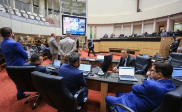 Alesc muda horários de votação devido ao ano eleitoral A imagem mostra o interior do Plenário Deputado Osni Régis na Assembleia Legislativa de Santa Catarina (Alesc), com carpete vermelho e as galerias superiores vazias. É uma foto do ambiente em que ocorrem as sessões legislativas descritas na matéria. No primeiro plano, deputados estaduais vestindo ternos estão sentados em bancadas equipadas com laptops (como Dell e Lenovo), focados em seus trabalhos, o que ilustra os parlamentares envolvidos nas discussões e votações da ordem do dia. Um homem de terno azul está em pé no canto esquerdo. Ao centro, uma grande tela de projeção suspensa exibe um close de um orador em um púlpito discursando, com o relógio de tempo de fala visível (00:09:34), indicando que a sessão deliberativa está em andamento. No fundo, a Mesa Diretora está reunida em uma plataforma elevada, e as bandeiras do Brasil e de Santa Catarina estão posicionadas ao centro, ladeadas por outros parlamentares. A cena captura o ambiente de trabalho e o rito legislativo que caracteriza o início do calendário especial de sessões antecipadas devido ao ano eleitoral, exemplificando uma sessão matinal deliberativa.