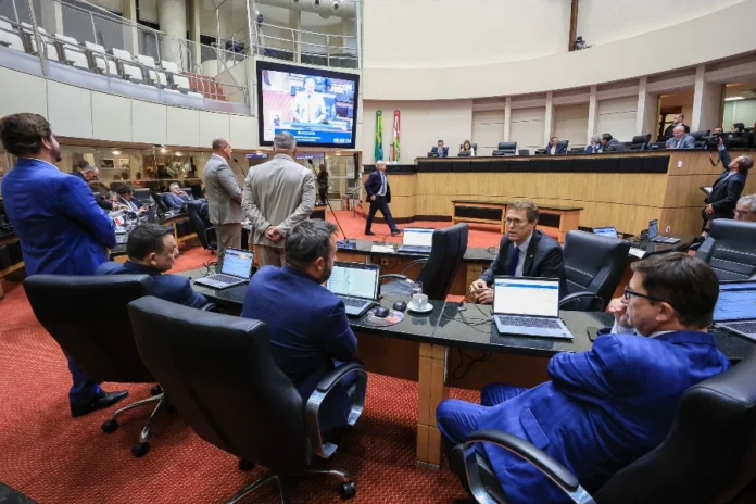 Alesc muda horários de votação devido ao ano eleitoral A imagem mostra o interior do Plenário Deputado Osni Régis na Assembleia Legislativa de Santa Catarina (Alesc), com carpete vermelho e as galerias superiores vazias. É uma foto do ambiente em que ocorrem as sessões legislativas descritas na matéria. No primeiro plano, deputados estaduais vestindo ternos estão sentados em bancadas equipadas com laptops (como Dell e Lenovo), focados em seus trabalhos, o que ilustra os parlamentares envolvidos nas discussões e votações da ordem do dia. Um homem de terno azul está em pé no canto esquerdo. Ao centro, uma grande tela de projeção suspensa exibe um close de um orador em um púlpito discursando, com o relógio de tempo de fala visível (00:09:34), indicando que a sessão deliberativa está em andamento. No fundo, a Mesa Diretora está reunida em uma plataforma elevada, e as bandeiras do Brasil e de Santa Catarina estão posicionadas ao centro, ladeadas por outros parlamentares. A cena captura o ambiente de trabalho e o rito legislativo que caracteriza o início do calendário especial de sessões antecipadas devido ao ano eleitoral, exemplificando uma sessão matinal deliberativa.