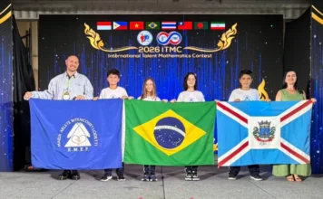 Içara recebe estudantes medalhistas em torneio mundial Uma fotografia colorida de seis pessoas em um palco durante uma cerimônia de premiação. Ao centro, quatro estudantes (duas meninas e dois meninos) vestindo camisetas brancas seguram, lado a lado, três bandeiras: a bandeira azul da escola E.M.E.F. Maria Arlete Bitencourt Lodetti, a bandeira do Brasil e a bandeira do município de Içara. À esquerda, um homem sorridente de camisa clara auxilia a segurar a bandeira da escola, e à direita, uma mulher em um vestido verde auxilia com a bandeira de Içara. Ao fundo, um telão digital exibe a logomarca do "2026 ITMC - International Talent Mathematics Contest" com as bandeiras dos países participantes no topo.