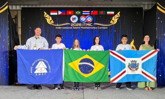 Alunos de Içara vencem mundial de matemática na Tailândia Uma fotografia colorida de seis pessoas em um palco durante uma cerimônia de premiação. Ao centro, quatro estudantes (duas meninas e dois meninos) vestindo camisetas brancas seguram, lado a lado, três bandeiras: a bandeira azul da escola E.M.E.F. Maria Arlete Bitencourt Lodetti, a bandeira do Brasil e a bandeira do município de Içara. À esquerda, um homem sorridente de camisa clara auxilia a segurar a bandeira da escola, e à direita, uma mulher em um vestido verde auxilia com a bandeira de Içara. Ao fundo, um telão digital exibe a logomarca do "2026 ITMC - International Talent Mathematics Contest" com as bandeiras dos países participantes no topo.