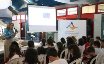 Alunos do Colégio Unesc participam de monitoramento de praias "Estudantes do Colégio Unesc assistem a uma palestra sobre o Projeto de Monitoramento de Praias (PMP-BP) no Museu de Zoologia da universidade. Uma palestrante loira em camiseta azul claro da Unesc fala em um microfone em frente a uma tela de projeção com o logo da IBAMA e um poster com o texto 'Projeto de Monitoramento de Praias'. Crianças em uniformes do Colégio Unesc estão sentadas em cadeiras brancas, ouvindo atentamente e fazendo anotações. O ambiente exibe modelos de animais marinhos pendurados no teto e um esqueleto de baleia atrás da palestrante."
