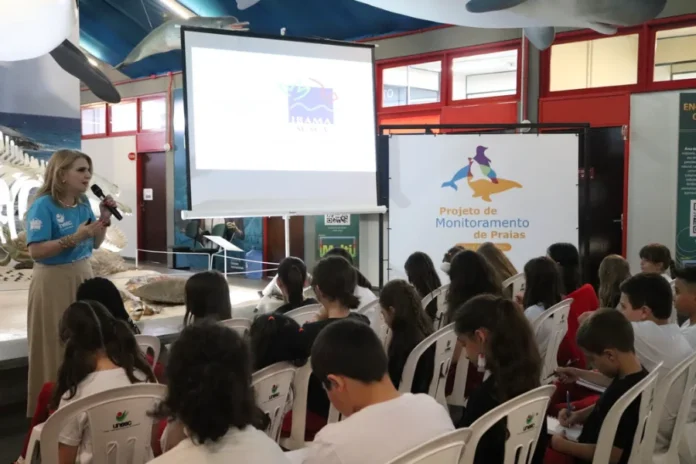 Alunos do Colégio Unesc participam de monitoramento de praias "Estudantes do Colégio Unesc assistem a uma palestra sobre o Projeto de Monitoramento de Praias (PMP-BP) no Museu de Zoologia da universidade. Uma palestrante loira em camiseta azul claro da Unesc fala em um microfone em frente a uma tela de projeção com o logo da IBAMA e um poster com o texto 'Projeto de Monitoramento de Praias'. Crianças em uniformes do Colégio Unesc estão sentadas em cadeiras brancas, ouvindo atentamente e fazendo anotações. O ambiente exibe modelos de animais marinhos pendurados no teto e um esqueleto de baleia atrás da palestrante."