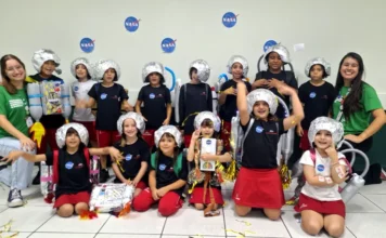Alunos do Colégio Unesc simulam viagem ao Sistema Solar Uma fotografia colorida em grupo de aproximadamente 15 crianças do ensino fundamental e duas jovens professoras, todas sorrindo em uma sala de atividades. As crianças estão vestidas com fantasias caseiras de astronautas, que incluem capacetes prateados feitos de papel alumínio e mochilas de materiais recicláveis (garrafas plásticas e caixas), algumas decoradas com o logotipo da NASA. Várias crianças usam camisetas pretas com um pequeno logotipo da NASA. Duas crianças no centro usam mochilas de foguete mais elaboradas. O grupo está posando em pé e ajoelhado no chão de cerâmica, com serpentinas e confetes dourados espalhados ao redor. Duas professoras, vestindo camisetas verdes idênticas, estão posicionadas nas extremidades do grupo. O fundo é uma parede branca decorada com vários adesivos grandes do logotipo circular azul e vermelho da NASA.