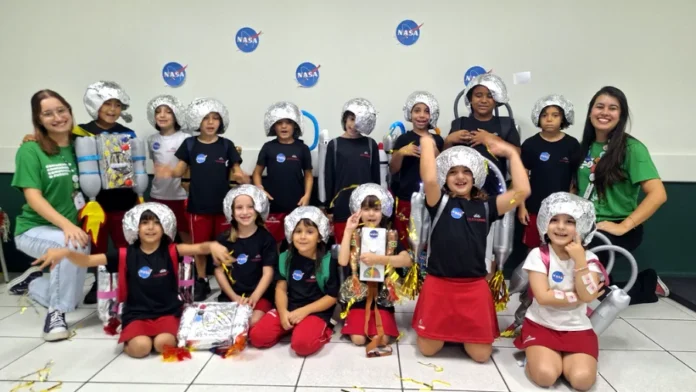 Alunos do Colégio Unesc simulam viagem ao Sistema Solar Uma fotografia colorida em grupo de aproximadamente 15 crianças do ensino fundamental e duas jovens professoras, todas sorrindo em uma sala de atividades. As crianças estão vestidas com fantasias caseiras de astronautas, que incluem capacetes prateados feitos de papel alumínio e mochilas de materiais recicláveis (garrafas plásticas e caixas), algumas decoradas com o logotipo da NASA. Várias crianças usam camisetas pretas com um pequeno logotipo da NASA. Duas crianças no centro usam mochilas de foguete mais elaboradas. O grupo está posando em pé e ajoelhado no chão de cerâmica, com serpentinas e confetes dourados espalhados ao redor. Duas professoras, vestindo camisetas verdes idênticas, estão posicionadas nas extremidades do grupo. O fundo é uma parede branca decorada com vários adesivos grandes do logotipo circular azul e vermelho da NASA.