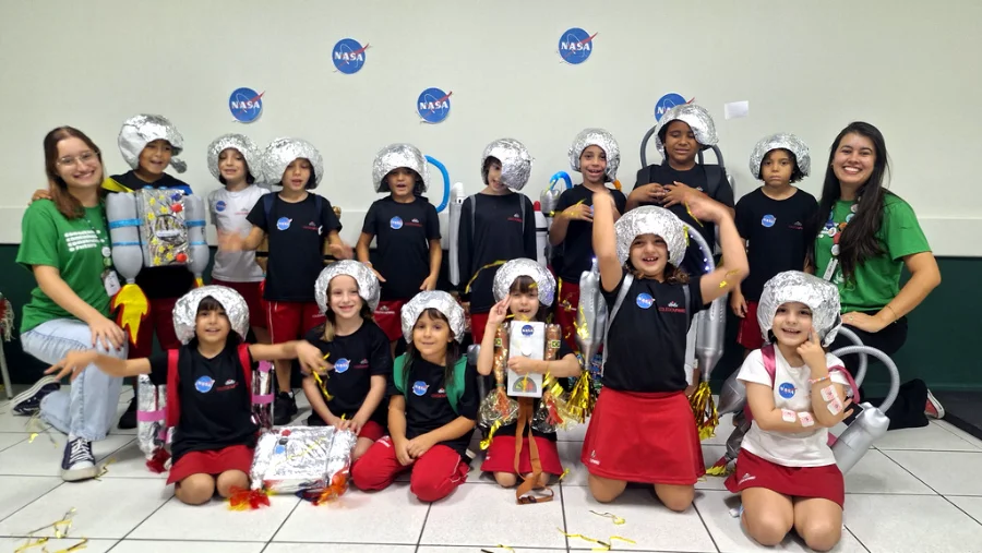 Uma fotografia colorida em grupo de aproximadamente 15 crianças do ensino fundamental e duas jovens professoras, todas sorrindo em uma sala de atividades. As crianças estão vestidas com fantasias caseiras de astronautas, que incluem capacetes prateados feitos de papel alumínio e mochilas de materiais recicláveis (garrafas plásticas e caixas), algumas decoradas com o logotipo da NASA. Várias crianças usam camisetas pretas com um pequeno logotipo da NASA. Duas crianças no centro usam mochilas de foguete mais elaboradas. O grupo está posando em pé e ajoelhado no chão de cerâmica, com serpentinas e confetes dourados espalhados ao redor. Duas professoras, vestindo camisetas verdes idênticas, estão posicionadas nas extremidades do grupo. O fundo é uma parede branca decorada com vários adesivos grandes do logotipo circular azul e vermelho da NASA.