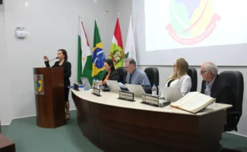 Ana Lucia da Silva toma posse na Câmara de Forquilhinha Uma foto colorida da 4ª Sessão Ordinária de 2026 da Câmara Municipal de Forquilhinha. Uma mulher com blazer preto gesticula enquanto fala em um púlpito de madeira à esquerda, prestando o compromisso legal para sua posse, conforme descrito no texto. Sentados à mesa semicircular ao centro e à direita estão quatro membros. As placas de identificação visíveis na mesa dizem 'IVONE MINATO' e 'JOSÉ NARDI PEREIRA', este último posicionado próximo a uma grande Bíblia aberta sobre a mesa. Ao fundo, bandeiras e um telão exibem o brasão da Câmara de Forquilhinha com a frase 'O PODER UNIDO É MAIS FORTE'.
