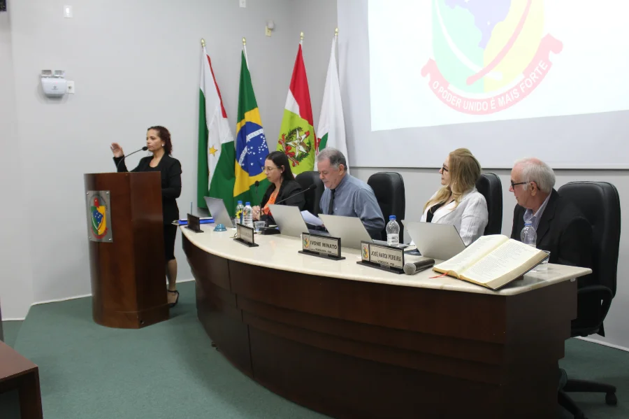 Uma foto colorida da 4ª Sessão Ordinária de 2026 da Câmara Municipal de Forquilhinha. Uma mulher com blazer preto gesticula enquanto fala em um púlpito de madeira à esquerda, prestando o compromisso legal para sua posse, conforme descrito no texto. Sentados à mesa semicircular ao centro e à direita estão quatro membros. As placas de identificação visíveis na mesa dizem 'IVONE MINATO' e 'JOSÉ NARDI PEREIRA', este último posicionado próximo a uma grande Bíblia aberta sobre a mesa. Ao fundo, bandeiras e um telão exibem o brasão da Câmara de Forquilhinha com a frase 'O PODER UNIDO É MAIS FORTE'.