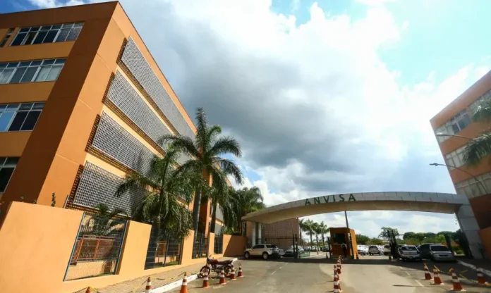 Anvisa aprova novos medicamentos para diabetes e câncer Fotografia externa do complexo de edifícios da sede da ANVISA, tirada de um ângulo baixo sob um céu dramaticamente nublado. À esquerda, um grande edifício de cor terracota com painéis de grelha metálica perfurada e palmeiras ao pé. Uma passarela curva elevada com o nome "ANVISA" em grandes letras metálicas no topo se estende entre o edifício esquerdo e outro bloco à direita. No primeiro plano, uma rua de acesso com carros estacionados, incluindo uma picape branca e uma motocicleta preta, está ladeada por cones de trânsito vermelhos e brancos.