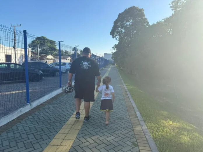 Araranguá SAMAE amplia vigilância na trilha do Belinzoni Um homem e uma criança caminham de mãos dadas, de costas para a câmera, pela trilha pavimentada do Parque Belinzoni, em Araranguá. O caminho é composto por blocos de concreto cinza com uma faixa central de piso tátil amarelo. À esquerda, há um cercado de grade azul que separa a trilha de um estacionamento com carros; à direita, observa-se uma área de gramado e árvores sob a luz intensa do sol. A cena ilustra o uso do espaço público para momentos de lazer em família.
