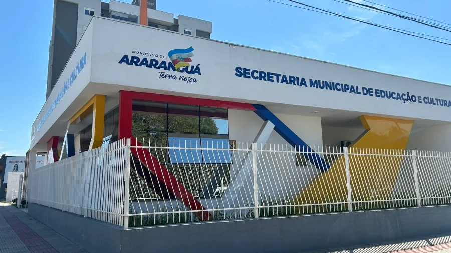Fachada lateral do prédio da Secretaria Municipal de Educação e Cultura de Araranguá, Santa Catarina. A edificação branca possui arquitetura moderna com detalhes geométricos em vermelho, amarelo e azul, exibindo o logotipo oficial do Município de Araranguá.