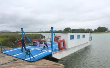 Araranguá suspende balsa para anúncio de nova ponte Vista lateral da balsa de Araranguá, com o nome 'MORRO DOS CONVENTOS' visível na lateral branca, atracada em uma rampa de concreto. Dois tripulantes são vistos no convés azul. O rio e a vegetação da margem oposta sob céu nublado formam o fundo, representando a travessia temporariamente suspensa para o evento da quarta ponte.