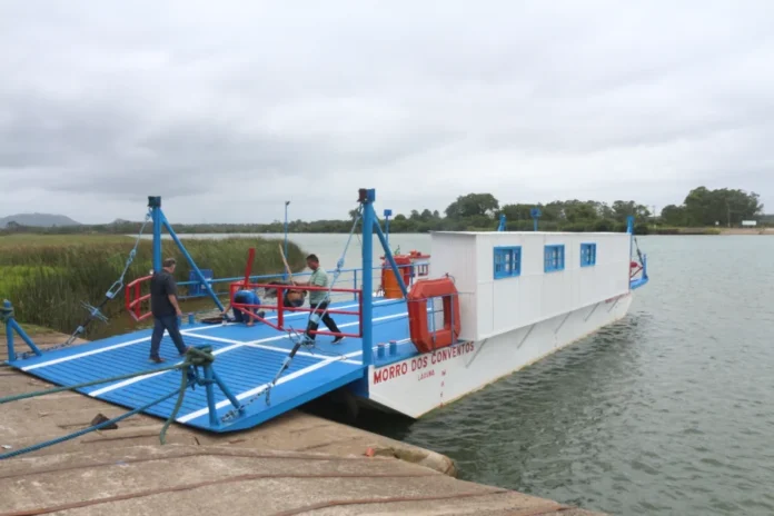 Araranguá suspende balsa para anúncio de nova ponte Vista lateral da balsa de Araranguá, com o nome 'MORRO DOS CONVENTOS' visível na lateral branca, atracada em uma rampa de concreto. Dois tripulantes são vistos no convés azul. O rio e a vegetação da margem oposta sob céu nublado formam o fundo, representando a travessia temporariamente suspensa para o evento da quarta ponte.