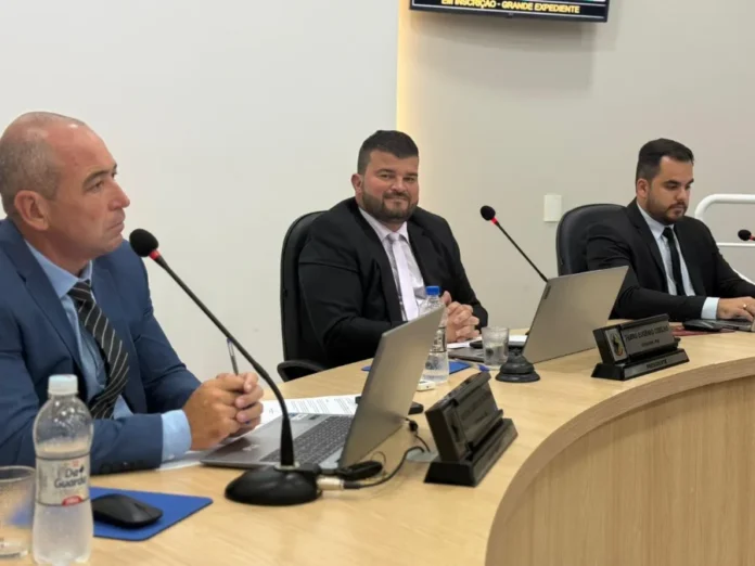 Arroio do Silva aprova REFIS 2026 e abafadores para autistas Uma fotografia colorida mostrando três homens sentados lado a lado em uma mesa de conferência curva de madeira clara em uma sala de reunião oficial. Da esquerda para a direita: um homem de meia-idade, careca, vestindo um terno azul-claro com gravata listrada azul e cinza, está sentado olhando para a frente com as mãos entrelaçadas sobre a mesa; o homem central, com barba e cabelo curto, veste um terno preto com camisa branca e uma gravata clara (bege ou rosa-claro), olha diretamente para a câmera e sorri levemente, com as mãos também entrelaçadas; o terceiro homem, mais jovem, veste um terno preto e gravata preta e está com o rosto ligeiramente voltado para baixo, olhando para algo na mesa diante dele. Diante de cada um dos homens há laptops (abertos ou fechados), microfones de conferência pescoço de ganso e garrafas de água. Na mesa diante do terceiro homem, há uma placa de identificação preta com a inscrição legível: "PEDRO BARROS COELHO, PRESIDENTE". O fundo é uma parede branca e clean, com um monitor ou painel de vídeo desfocado na parte superior central, exibindo texto ilegível. A iluminação é de escritório, difusa e uniforme.