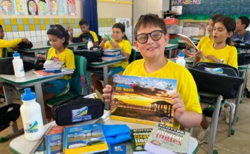 Arroio do Silva entrega kits escolares para 2.400 alunos Aluno da rede municipal de Balneário Arroio do Silva sorri em sala de aula ao lado de kit de material escolar personalizado com cadernos, estojo, dicionários e garrafa de água sobre a mesa.