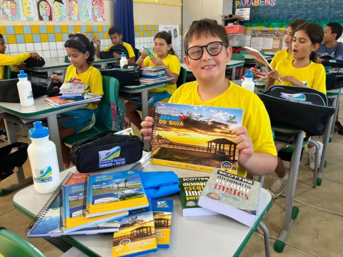 Arroio do Silva entrega kits escolares para 2.400 alunos Aluno da rede municipal de Balneário Arroio do Silva sorri em sala de aula ao lado de kit de material escolar personalizado com cadernos, estojo, dicionários e garrafa de água sobre a mesa.