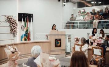 Arroio do Silva premia 11 mulheres com troféu especial Uma mulher orando ao microfone em um púlpito de madeira na Câmara de Vereadores de Balneário Arroio do Silva durante a Sessão Solene do Troféu Lucília da Silva Borges, com a galeria de público ao fundo e bandeiras oficiais ao lado.