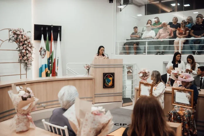 Arroio do Silva premia 11 mulheres com troféu especial (1) Uma mulher orando ao microfone em um púlpito de madeira na Câmara de Vereadores de Balneário Arroio do Silva durante a Sessão Solene do Troféu Lucília da Silva Borges, com a galeria de público ao fundo e bandeiras oficiais ao lado.
