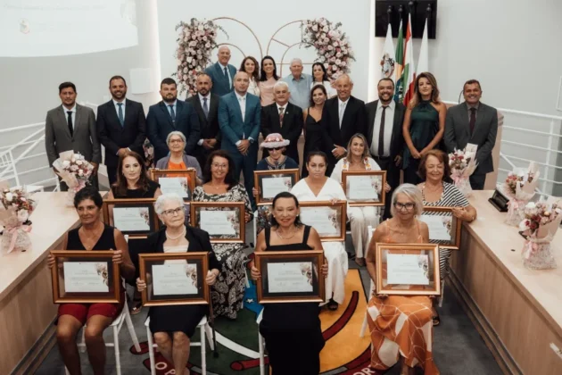 Foto em grupo das 11 mulheres homenageadas com o Trof&eacute;u Luc&iacute;lia da Silva Borges, segurando seus certificados e flores, ao lado de vereadores e autoridades na C&acirc;mara de Vereadores de Balne&aacute;rio Arroio do Silva.