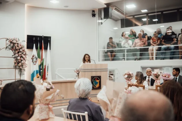 Sess&atilde;o Solene do Trof&eacute;u Luc&iacute;lia da Silva Borges na C&acirc;mara de Vereadores de Balne&aacute;rio Arroio do Silva, mostrando uma mulher discursando no p&uacute;lpito e uma das homenageadas com chap&eacute;u segurando seu certificado na plateia.