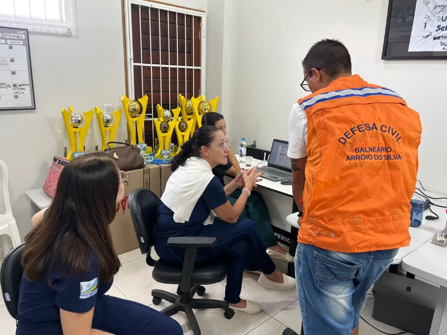 Três mulheres e um homem com colete laranja da Defesa Civil de Balneário Arroio do Silva estão reunidos em torno de uma mesa com um notebook. Eles realizam procedimentos técnicos de cadastro e organização, simulando o atendimento em situações de desastre. Ao fundo, em uma estante, é possível ver troféus esportivos amarelos.