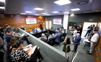CCJ aprova direitos de PcD para diabéticos tipo 1 em SC Fotografia panorâmica de uma sala de audiência formal durante uma reunião da Comissão de Constituição e Justiça (CCJ). Na área central, membros da comissão estão sentados em torno de uma mesa em formato de U, voltados para duas grandes telas de televisão que exibem, de forma clara, o texto 'COMISSÃO DE CONSTITUIÇÃO E JUSTIÇA'. No primeiro plano e à esquerda, a audiência está sentada em cadeiras em estilo de anfiteatro. Uma mulher de blusa estampada em preto e branco olha para o celular enquanto segura papéis. Várias outras pessoas, algumas em pé e outras sentadas, ocupam o espaço. O ambiente tem painéis de madeira nas paredes e iluminação embutida no teto.