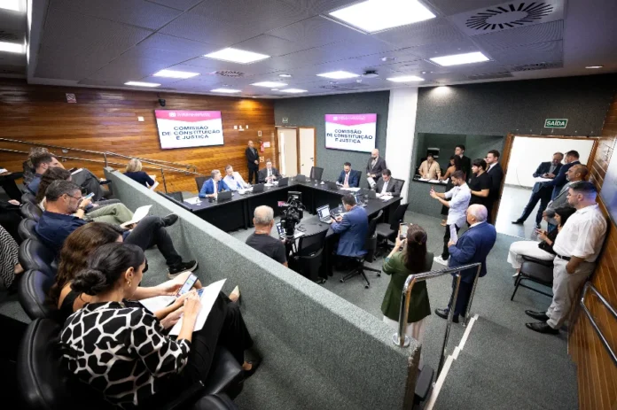 CCJ aprova direitos de PcD para diabéticos tipo 1 em SC Fotografia panorâmica de uma sala de audiência formal durante uma reunião da Comissão de Constituição e Justiça (CCJ). Na área central, membros da comissão estão sentados em torno de uma mesa em formato de U, voltados para duas grandes telas de televisão que exibem, de forma clara, o texto 'COMISSÃO DE CONSTITUIÇÃO E JUSTIÇA'. No primeiro plano e à esquerda, a audiência está sentada em cadeiras em estilo de anfiteatro. Uma mulher de blusa estampada em preto e branco olha para o celular enquanto segura papéis. Várias outras pessoas, algumas em pé e outras sentadas, ocupam o espaço. O ambiente tem painéis de madeira nas paredes e iluminação embutida no teto.
