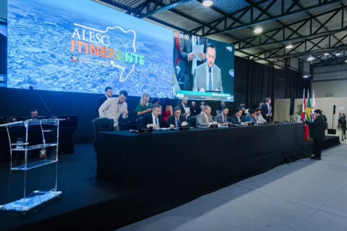 CCJ aprova reconhecimento do Tiro de Laço como esporte em SC Mesa diretora da ALESC Itinerante em Curitibanos, Santa Catarina, durante sessão da Comissão de Constituição e Justiça (CCJ). Deputados estaduais SC participam de reunião parlamentar com telão ao fundo exibindo o logotipo da Assembleia Legislativa.