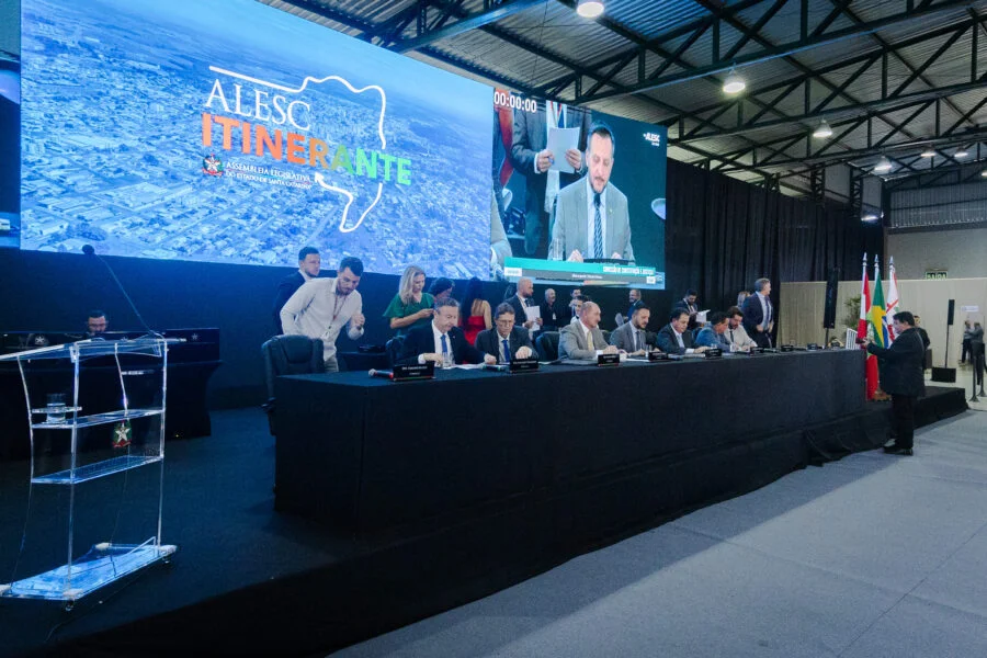 Mesa diretora da ALESC Itinerante em Curitibanos, Santa Catarina, durante sessão da Comissão de Constituição e Justiça (CCJ). Deputados estaduais SC participam de reunião parlamentar com telão ao fundo exibindo o logotipo da Assembleia Legislativa.
