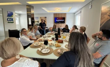 CDL Araranguá lança projeto Café com a Presidente Fotografia em plano médio de uma reunião realizada em ambiente moderno e bem iluminado. Cerca de dez pessoas, entre homens e mulheres, estão sentadas ao redor de uma grande mesa branca retangular, posta para um café da manhã com xícaras, garrafas térmicas, pães e bolos. Ao fundo, à esquerda, destaca-se o logotipo azul e amarelo da CDL Araranguá em uma pilastra branca. Um homem está de pé ao centro, conversando com o grupo, enquanto os demais acompanham a fala. No fundo da sala, uma televisão exibe a palavra "ARARANGUÁ".