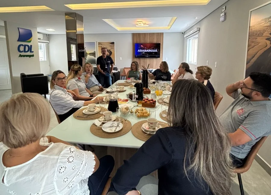 Fotografia em plano médio de uma reunião realizada em ambiente moderno e bem iluminado. Cerca de dez pessoas, entre homens e mulheres, estão sentadas ao redor de uma grande mesa branca retangular, posta para um café da manhã com xícaras, garrafas térmicas, pães e bolos. Ao fundo, à esquerda, destaca-se o logotipo azul e amarelo da CDL Araranguá em uma pilastra branca. Um homem está de pé ao centro, conversando com o grupo, enquanto os demais acompanham a fala. No fundo da sala, uma televisão exibe a palavra 