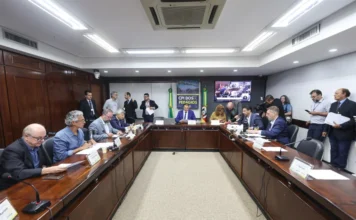 CPI dos Pedágios cobra explicações sobre editais no RS A imagem apresenta uma sala de audiências da Assembleia Legislativa em plano aberto. No centro, parlamentares e convidados estão sentados ao redor de uma grande mesa de madeira escura em formato de "U", equipada com microfones e documentos. Ao fundo, posiciona-se um banner vertical com a inscrição "CPI DOS PEDÁGIOS" e uma tela de transmissão simultânea que exibe o plenário. O ambiente possui iluminação clara e painéis de madeira nas paredes, caracterizando uma sessão oficial de debate parlamentar.