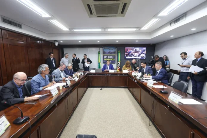 CPI dos Pedágios cobra explicações sobre editais no RS A imagem apresenta uma sala de audiências da Assembleia Legislativa em plano aberto. No centro, parlamentares e convidados estão sentados ao redor de uma grande mesa de madeira escura em formato de "U", equipada com microfones e documentos. Ao fundo, posiciona-se um banner vertical com a inscrição "CPI DOS PEDÁGIOS" e uma tela de transmissão simultânea que exibe o plenário. O ambiente possui iluminação clara e painéis de madeira nas paredes, caracterizando uma sessão oficial de debate parlamentar.