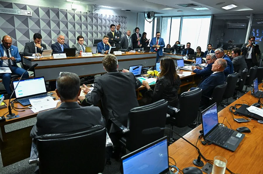 Vista geral de uma reunião de comissão parlamentar. Vários homens de terno estão sentados em uma grande mesa em forma de U com laptops. Uma pessoa de terno está em pé e falando no centro da mesa. Ao fundo, uma parede com um painel geométrico cinza e o logotipo do Senado Federal. Outras pessoas estão sentadas ou em pé ao redor.
