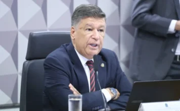 CPMI do INSS ouve presidente da Dataprev sobre fraudes Fotografia em plano médio do senador Carlos Viana, presidente da CPMI do INSS. Ele é um homem de pele clara, cabelos curtos e escuros, veste terno azul-marinho e gravata com listras vermelhas e pretas. O senador está sentado à mesa de reuniões, com os braços cruzados, falando ao microfone. À sua frente, há um notebook e um copo com água. O fundo apresenta uma parede com relevos geométricos em tons de cinza.