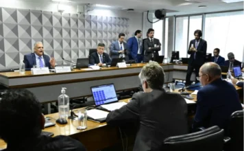 CPMI do INSS recebe dados sigilosos do banqueiro Vorcaro Foto colorida em ângulo médio que mostra uma sala de reuniões de comissão governamental em andamento. Em uma mesa principal de madeira, elevada e em destaque, um homem de terno azul e gravata azul-clara (orador principal) está sentado e fala ao microfone, com a mão esquerda levantada e a palma aberta para frente. Ao seu lado, outro homem de terno escuro e óculos está sentado, olhando para baixo para documentos. Atrás da mesa principal, vários homens em pé, de terno, observam a cena. Em primeiro plano, as costas de duas pessoas sentadas à mesa principal olhando para o orador. Sobre as mesas, há laptops, microfones de mesa, copos e garrafas de álcool em gel. A parede ao fundo tem um painel geométrico texturizado em tons de cinza. Janelas grandes dão para fora. O ambiente é formal e bem iluminado.