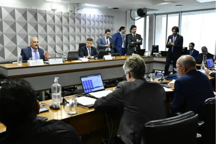 CPMI do INSS recebe dados sigilosos do banqueiro Vorcaro Foto colorida em ângulo médio que mostra uma sala de reuniões de comissão governamental em andamento. Em uma mesa principal de madeira, elevada e em destaque, um homem de terno azul e gravata azul-clara (orador principal) está sentado e fala ao microfone, com a mão esquerda levantada e a palma aberta para frente. Ao seu lado, outro homem de terno escuro e óculos está sentado, olhando para baixo para documentos. Atrás da mesa principal, vários homens em pé, de terno, observam a cena. Em primeiro plano, as costas de duas pessoas sentadas à mesa principal olhando para o orador. Sobre as mesas, há laptops, microfones de mesa, copos e garrafas de álcool em gel. A parede ao fundo tem um painel geométrico texturizado em tons de cinza. Janelas grandes dão para fora. O ambiente é formal e bem iluminado.