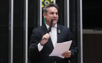 Câmara aprova novos critérios para indenização de armas Deputado Zucco discursa na Câmara dos Deputados, em Brasília, durante sessão de comissão. Ele segura papéis e um microfone, com o brasão do Brasil ao fundo. Imagem relacionada à aprovação do projeto de lei de indenização de armas.